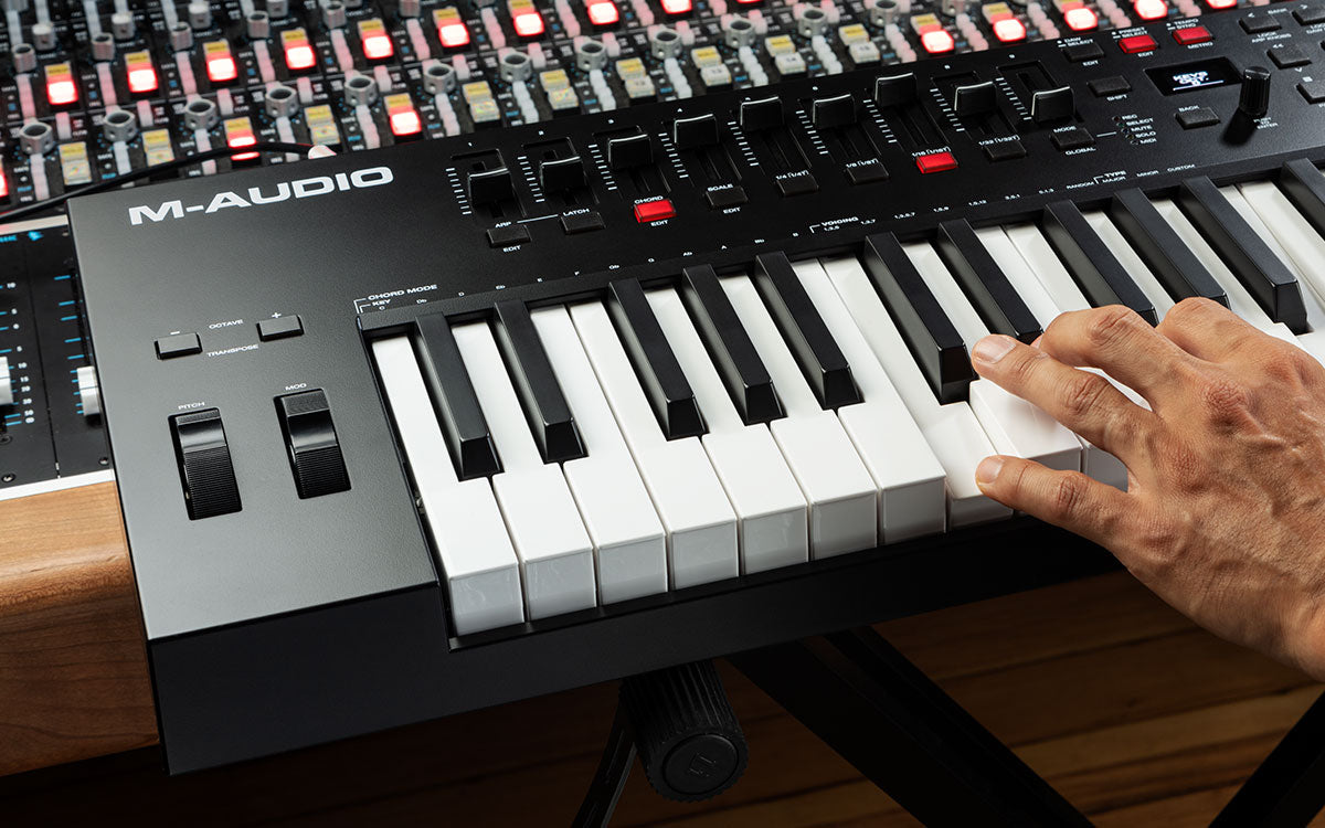 M-Audio keyboard controller with a hand on the keys, surrounded by audio equipment.