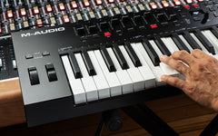 M-Audio keyboard controller with a hand on the keys, surrounded by audio equipment.